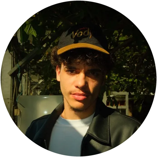 yeldee: curls, black cap, leather jacket, in the sun between plants.
